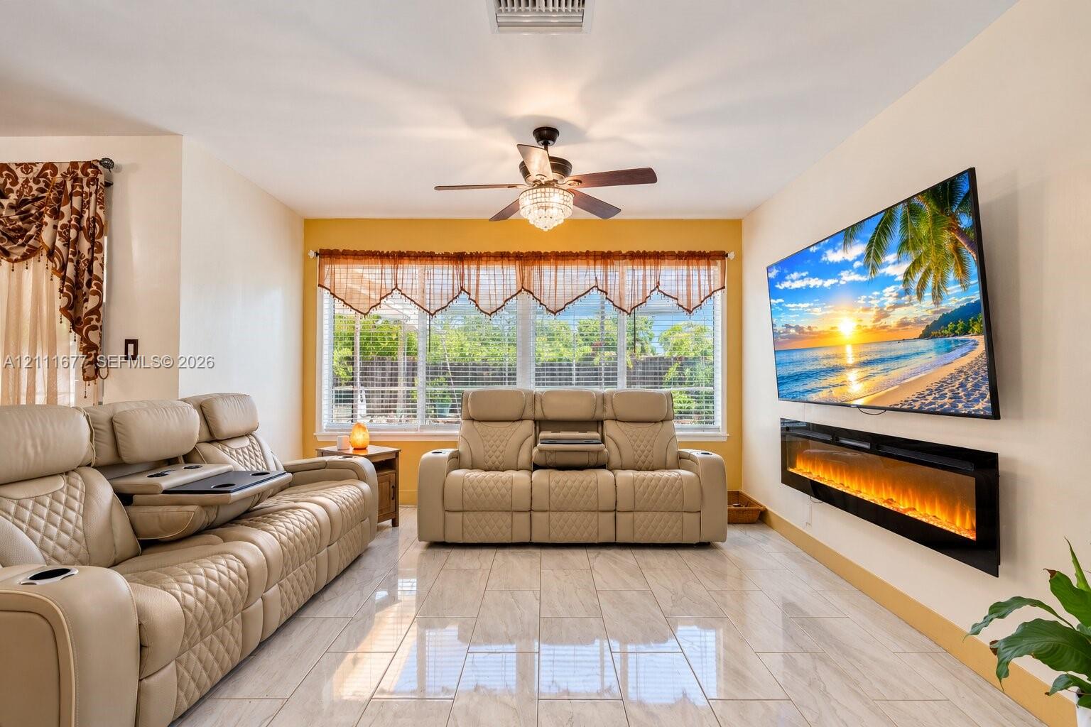 Living area featuring polished tile flooring, wall-mounted electric fireplace, ceiling fan with integrated lighting, and multiple windows