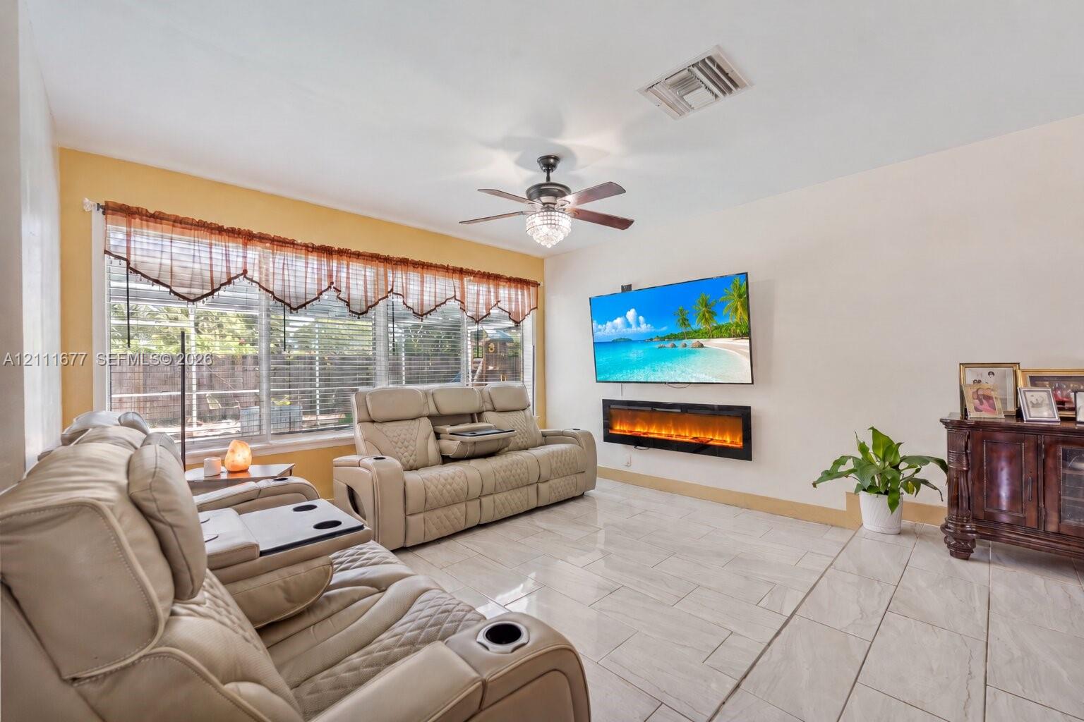 Living area featuring large format tile flooring, a wall-mounted electric fireplace, and a ceiling fan with integrated lighting