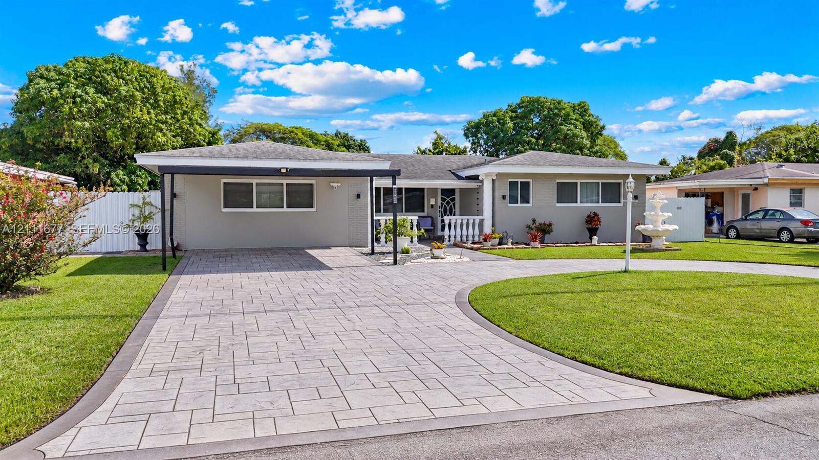 Single-story exterior featuring a paver driveway, covered carport, manicured lawn, tiered water fountain, and white balustrade railing Single-story exterior featuring a paver driveway, covered carport, manicured lawn, tiered water fountain, and white balustrade railing