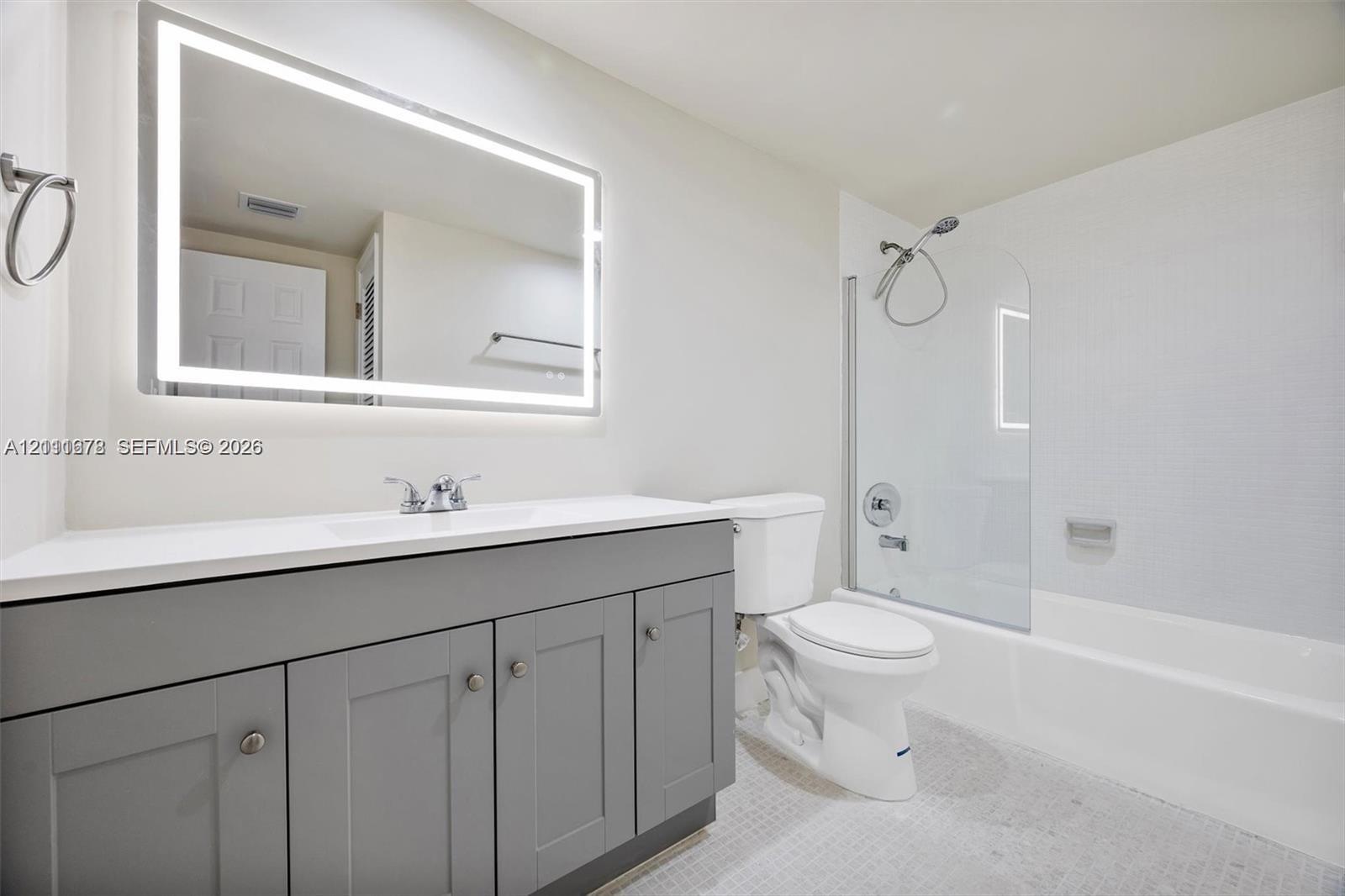Bathroom featuring a grey shaker-style vanity with a solid surface countertop, an illuminated rectangular wall mirror, a glass shower partition, and a built-in bathtub with a wall-mounted shower head