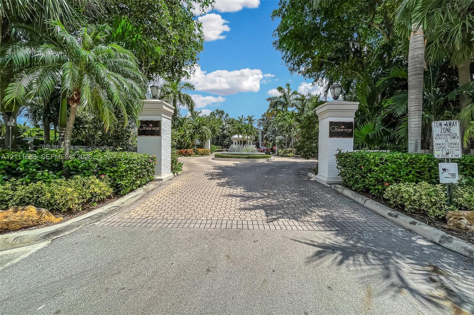Gated entrance featuring decorative stone pillars, paver driveway, mature palm trees, lush perimeter landscaping, and a central fountain Gated entrance featuring decorative stone pillars, paver driveway, mature palm trees, lush perimeter landscaping, and a central fountain