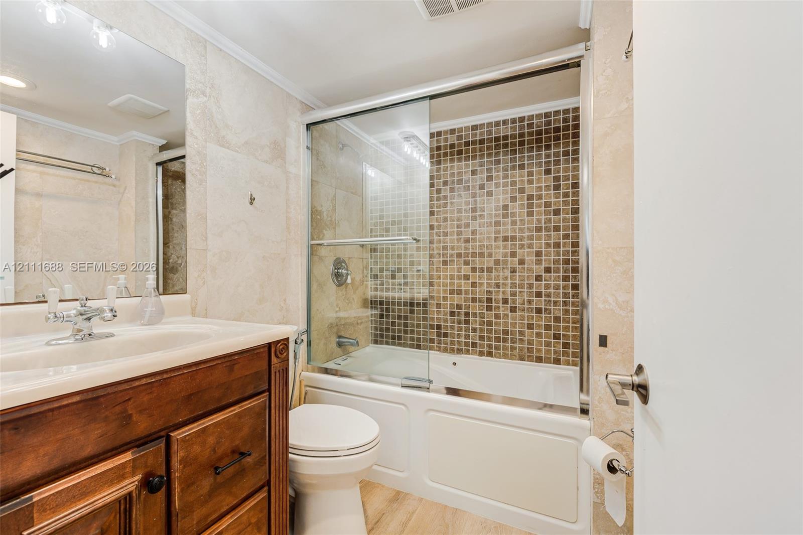 Bathroom featuring a wood-finish vanity with an integrated sink, large wall-mounted mirror, glass-enclosed shower and tub combination, and tiled surround