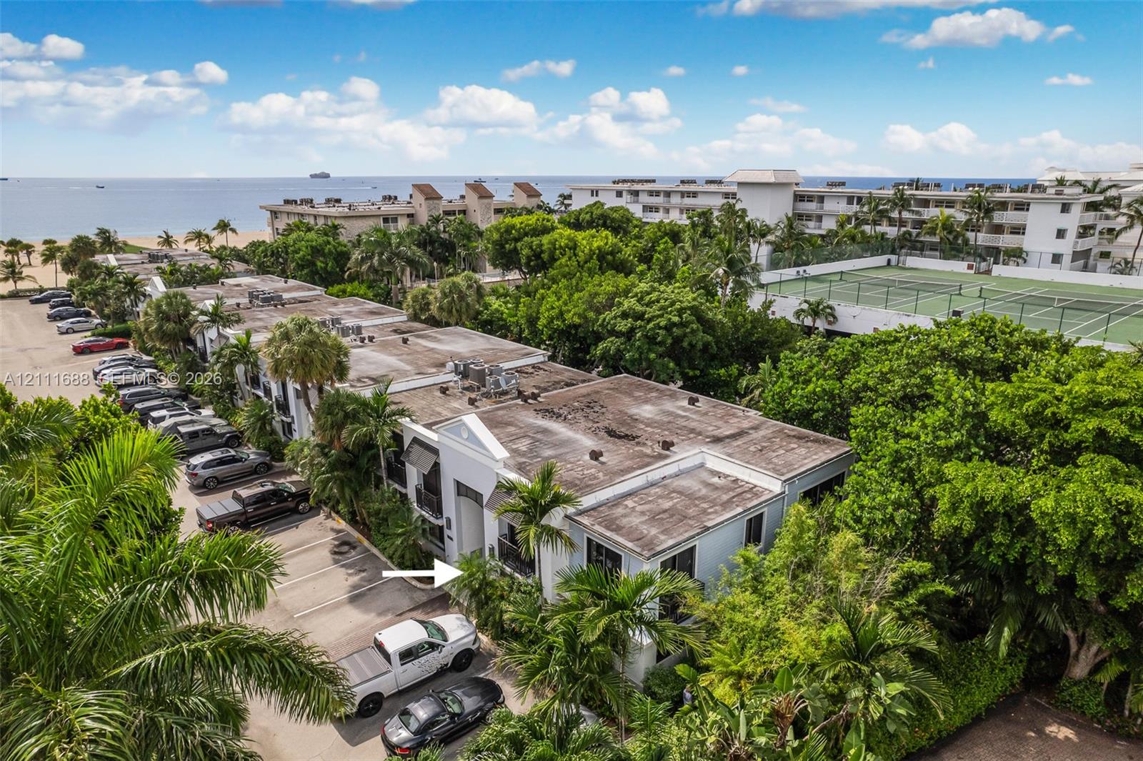 Aerial perspective of a multi-story residential building complex featuring white exterior facades, mature palm landscaping, on-site paved parking, and a fenced tennis court Aerial perspective of a multi-story residential building complex featuring white exterior facades, mature palm landscaping, on-site paved parking, and a fenced tennis court