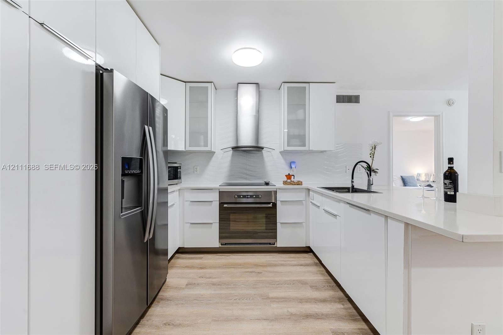 Kitchen featuring high-gloss white cabinetry, stainless steel appliances, a textured tile backsplash, wood-finish flooring, and a quartz countertop peninsula