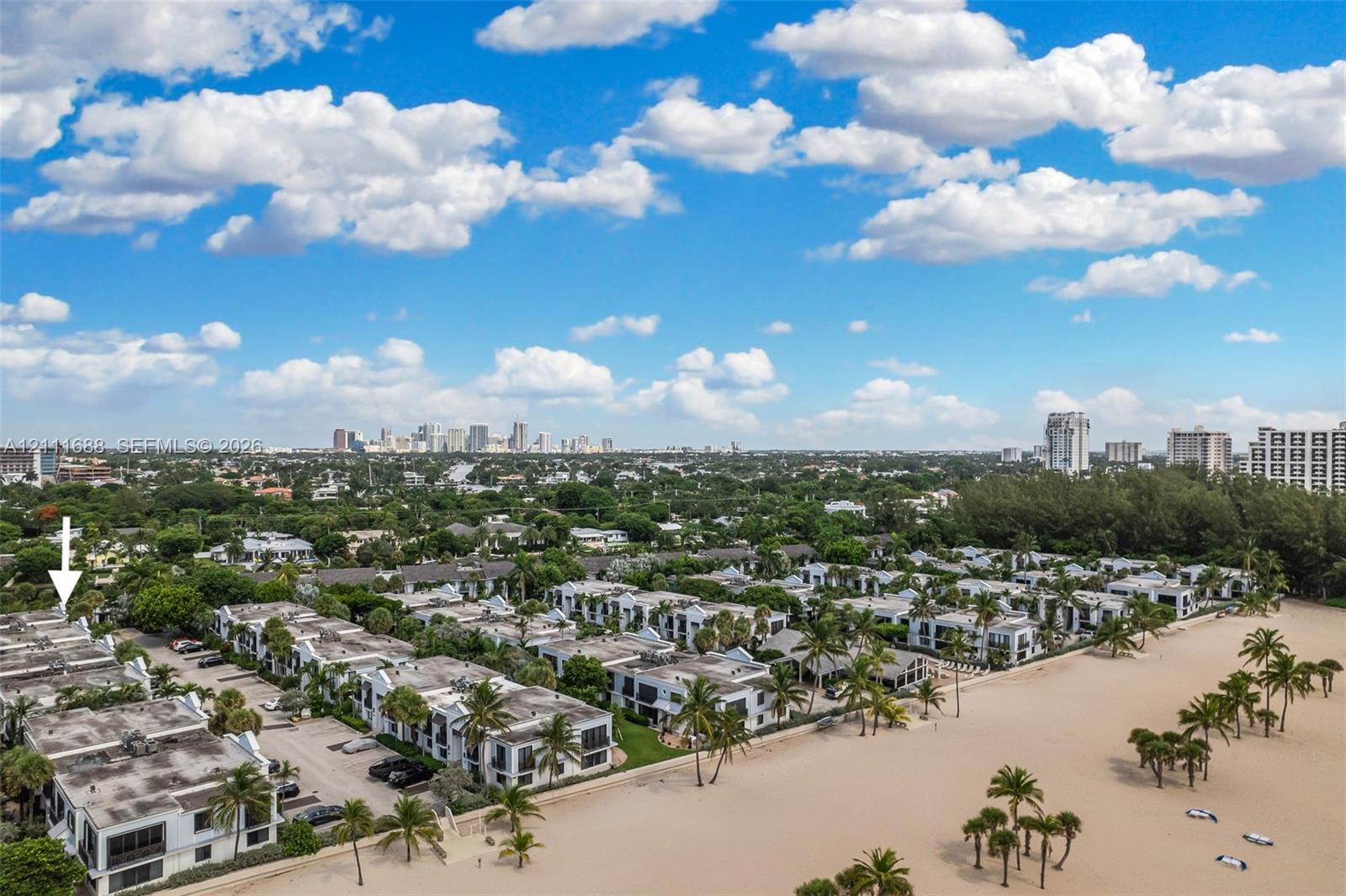 Aerial perspective of a multi-unit complex featuring direct beach access, coastal vegetation, and a cityscape horizon