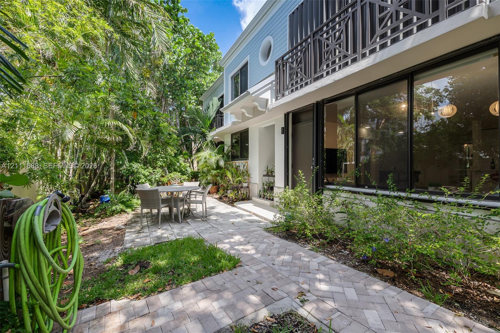 Exterior patio featuring herringbone-patterned brick pavers, two-story light blue siding, an upper-level balcony with geometric railing, and expansive floor-to-ceiling glass windows