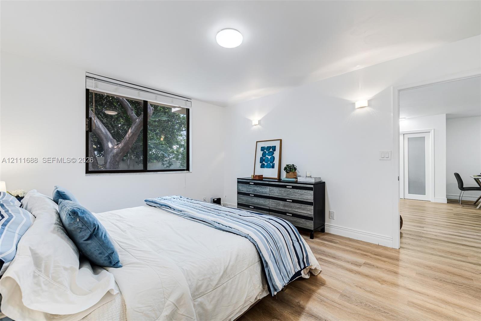 Neutral-toned interior featuring wood-finish flooring, dual wall-mounted light fixtures, a recessed ceiling light, and a large single-pane window