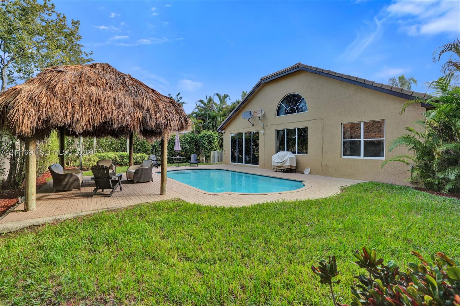 Stucco-exterior residence featuring an in-ground swimming pool, a thatched-roof tiki hut, a paved patio, and a manicured lawn