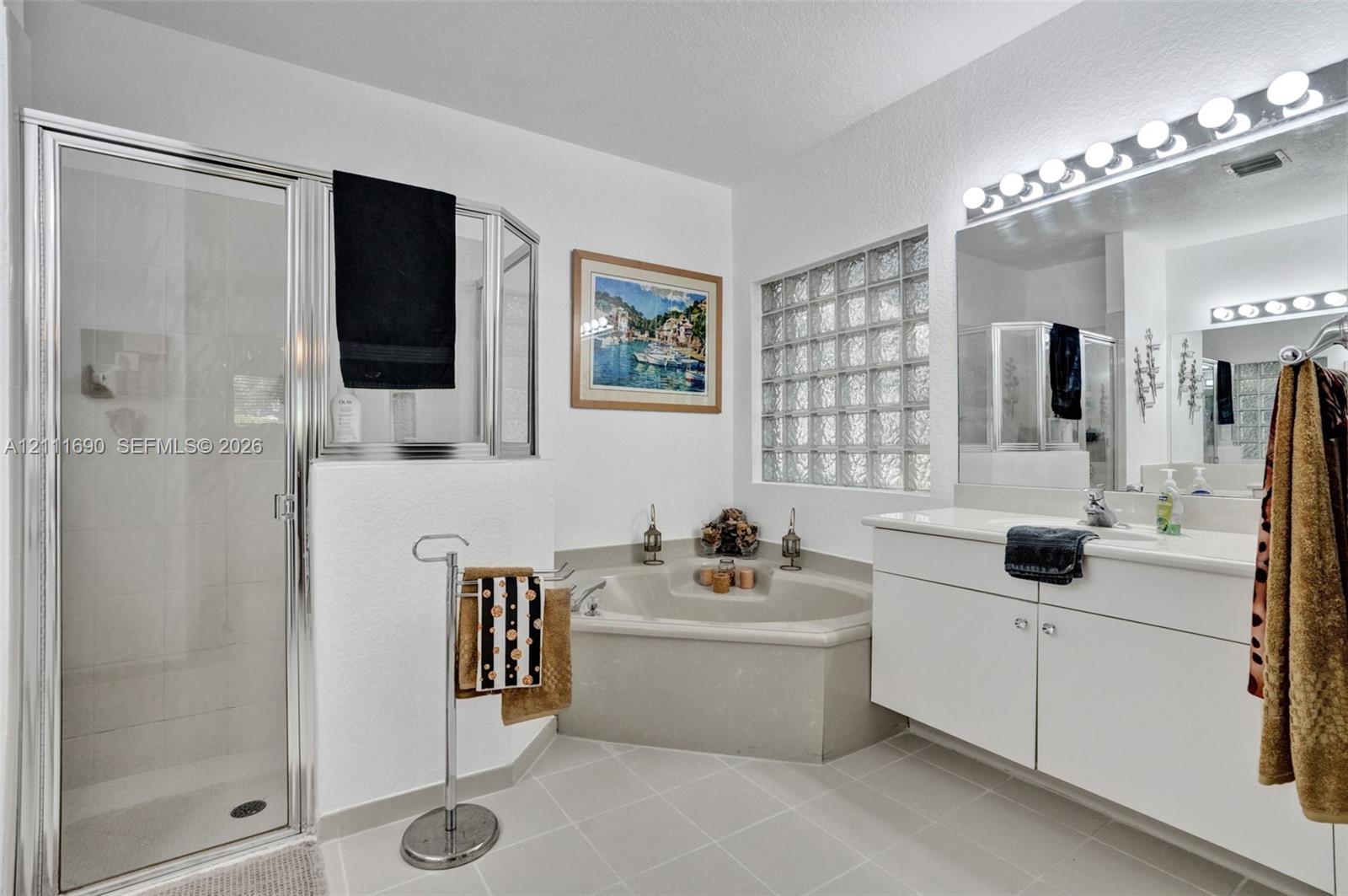 Bathroom featuring a walk-in shower with a glass enclosure, a corner soaking tub, a white vanity with integrated basin, glass block accent window, and a multi-bulb vanity light fixture