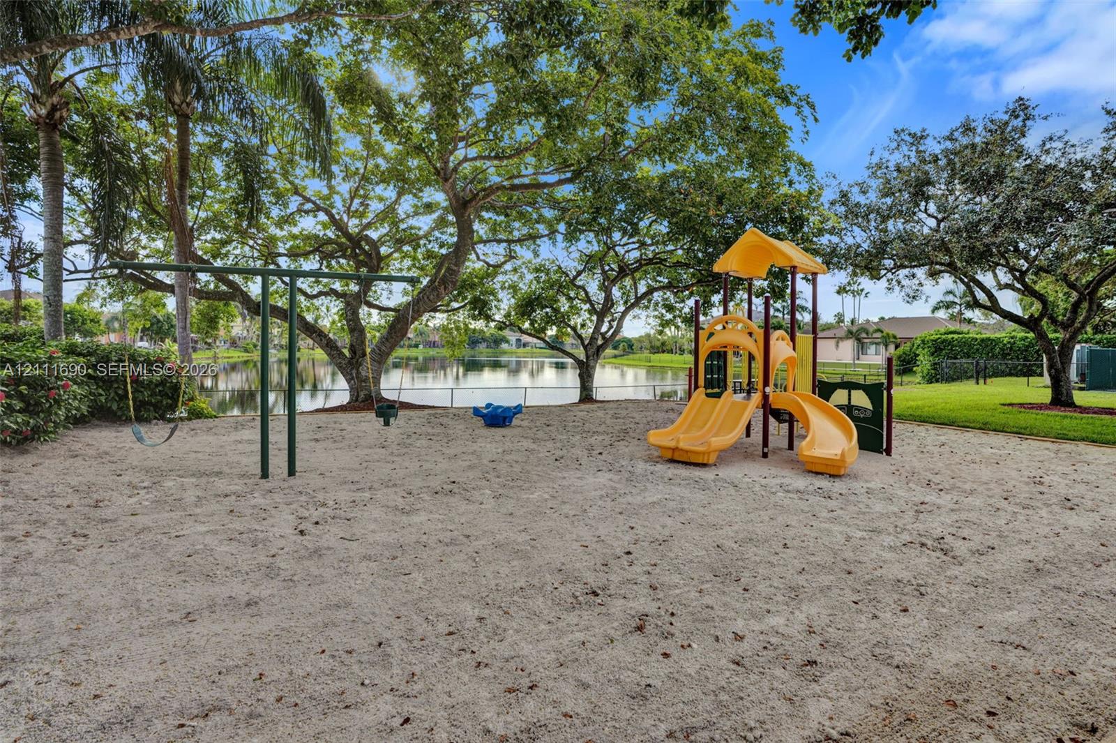 Outdoor recreation area featuring a sand-surface playground with a multi-slide structure and swing set, situated among mature trees near a pond