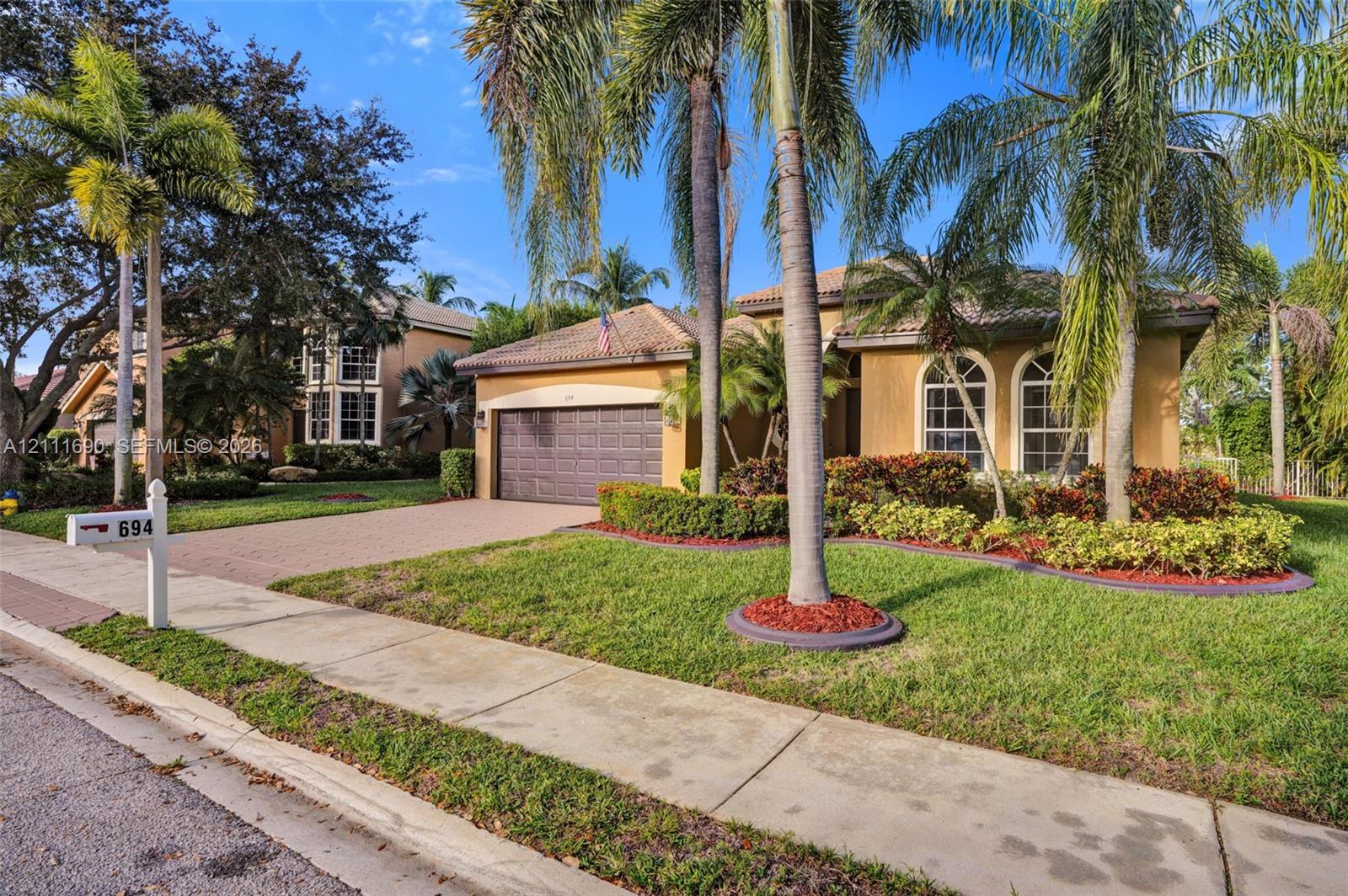 Single-story stucco exterior featuring a tiled roof, arched window detailing, an attached two-car garage, and manicured landscaping with mature palms