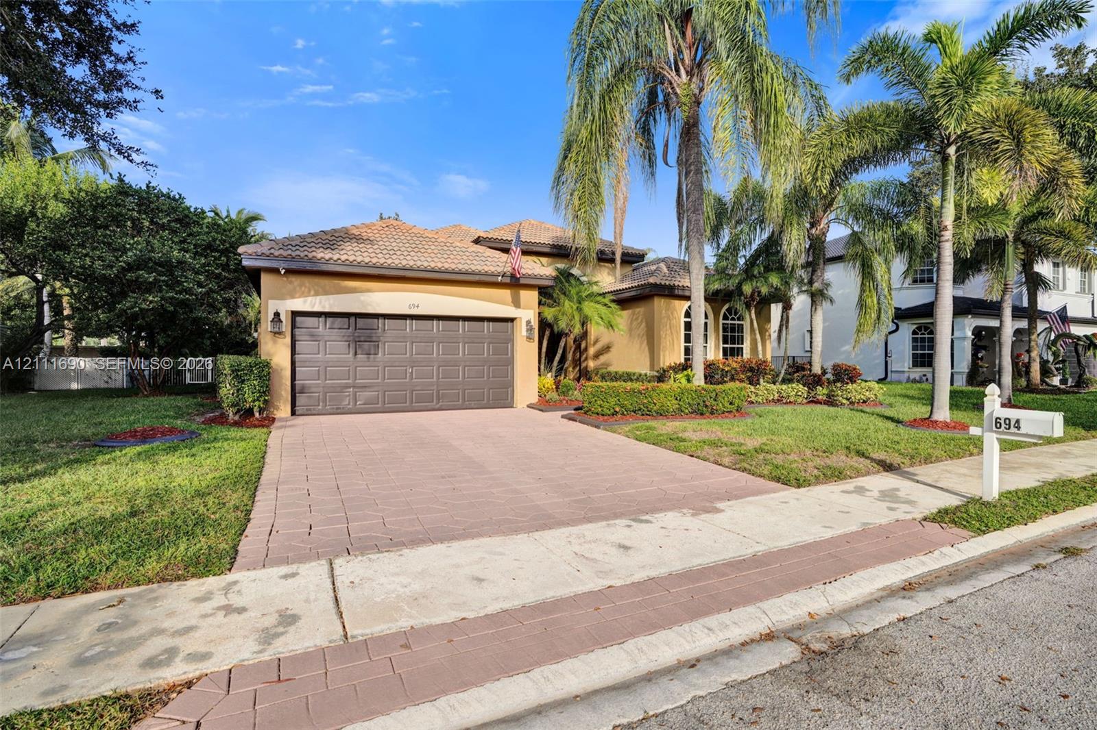 Stucco exterior featuring a multi-hipped tile roof, paved driveway, attached two-car garage, and established palm trees