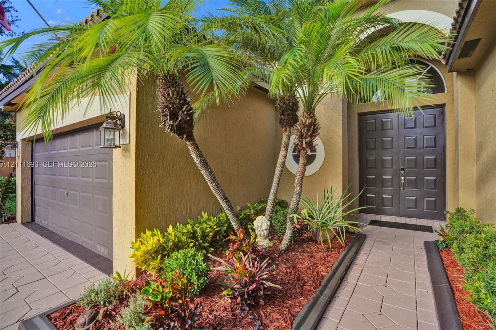 Stucco exterior featuring dual palm trees, a paver walkway, a raised-panel double front entry door, and a multi-section garage door