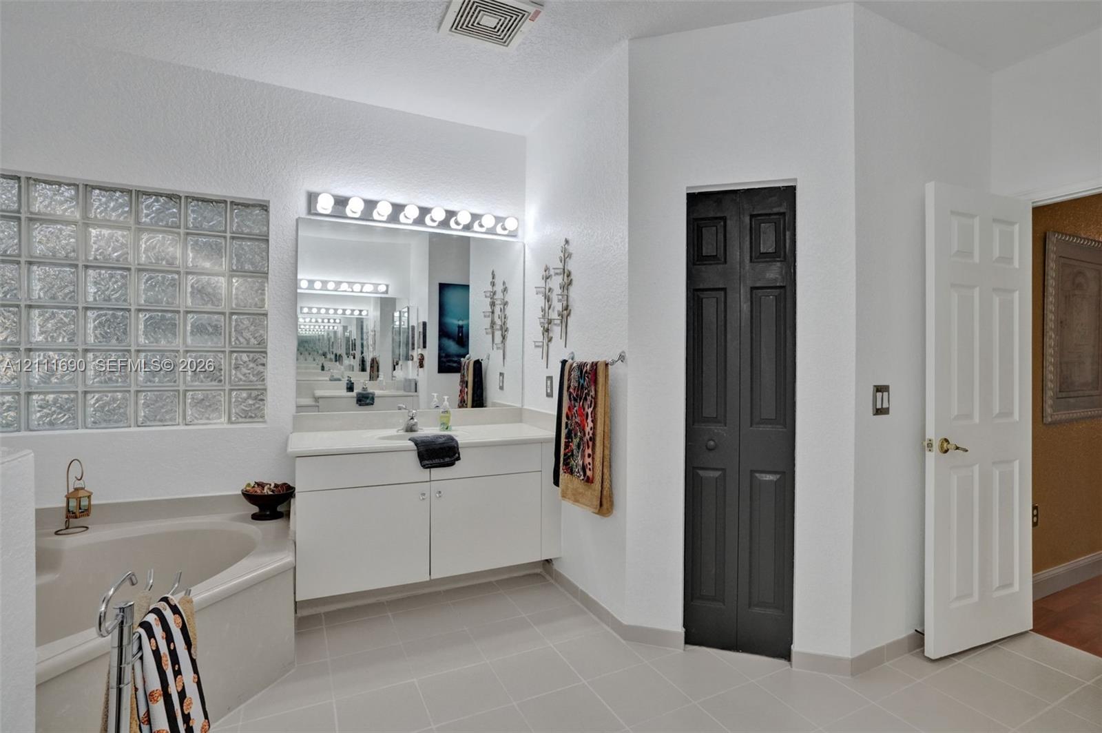 Bathroom featuring a glass block window, built-in soaking tub, white vanity with stone-surface countertop, full-width vanity mirror, and neutral tile flooring