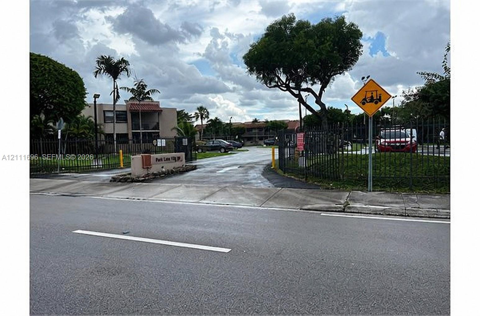 Gated community entrance featuring metal perimeter fencing, paved driveway access, and mature landscaping Gated community entrance featuring metal perimeter fencing, paved driveway access, and mature landscaping