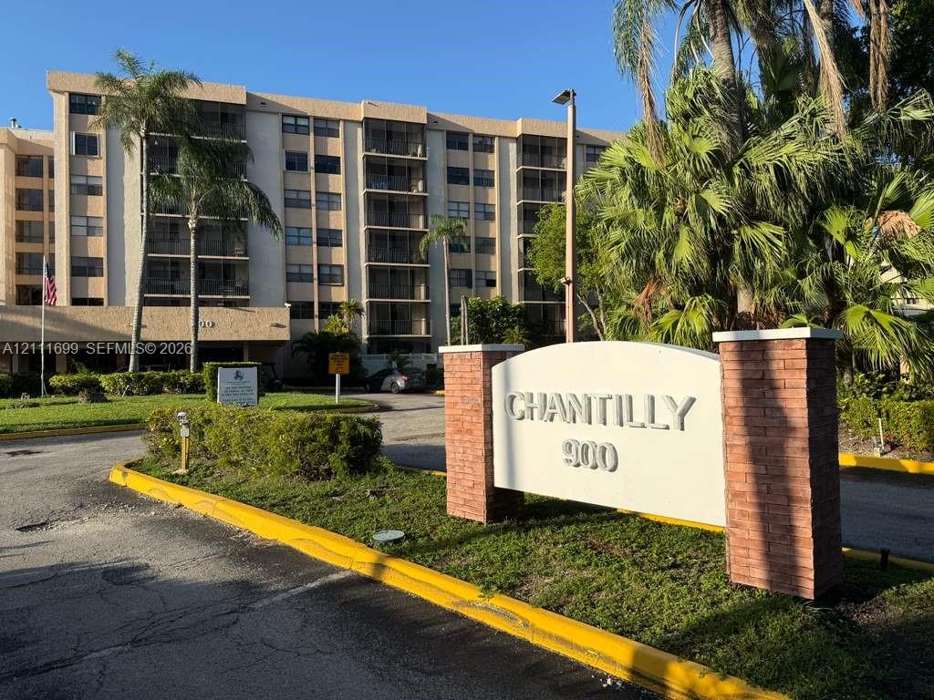 Multi-story residential building featuring covered balconies, brick-accented monument signage, paved entry drive, and mature tropical landscaping