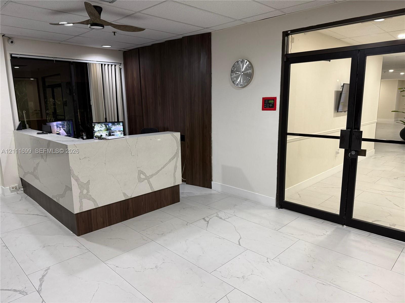 Reception area featuring a stone-topped built-in desk with wood-finish paneling, large-format tile flooring, a dark-finish ceiling fan, and double glass doors with black framing