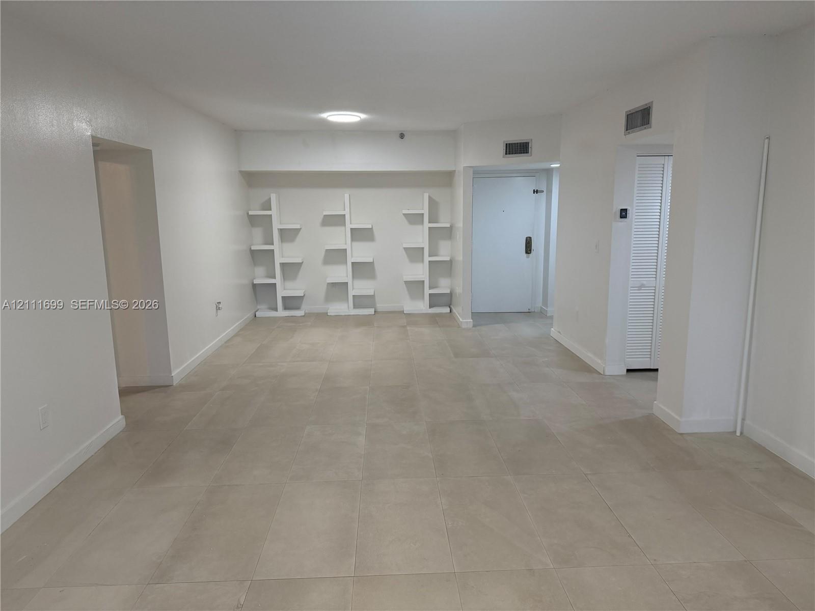 Neutral-toned floor tiles, three built-in white tiered shelving units, and a closet with louvered doors