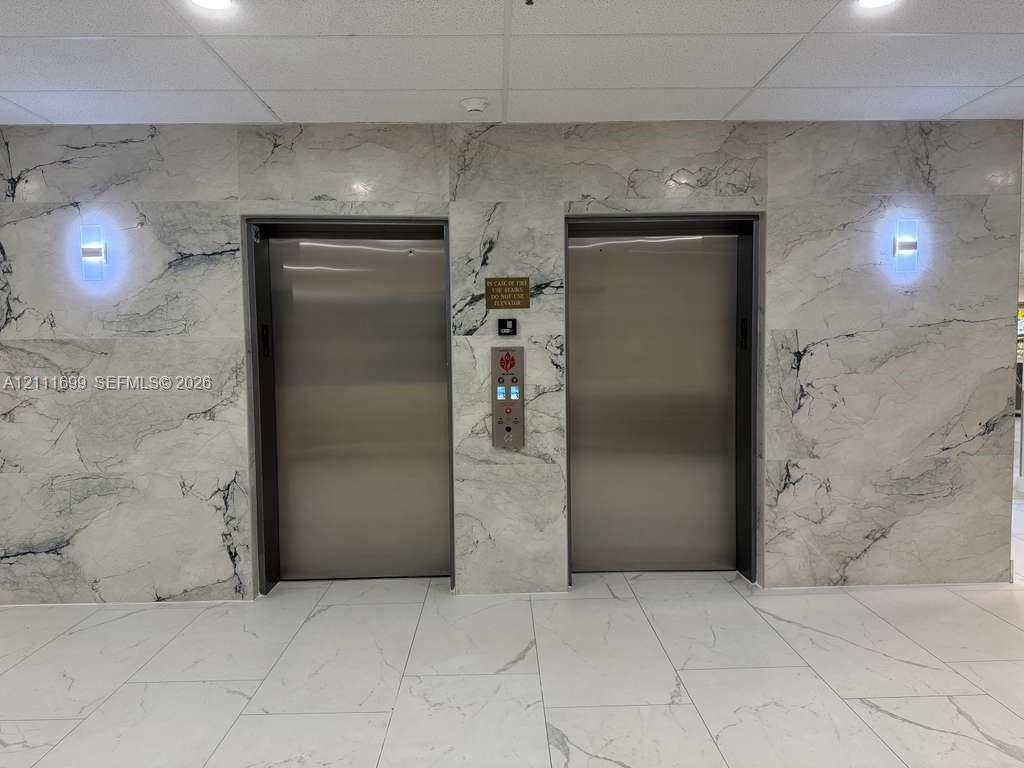 Dual stainless steel elevator doors featuring floor-to-ceiling stone-clad surround, large-format tile flooring, and wall-mounted light fixtures