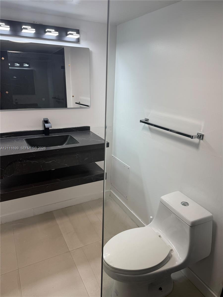 Floating vanity with a dark-toned stone surface and integrated basin, matte black faucet, wall-mounted rectangular mirror with integrated lighting, and one-piece white toilet