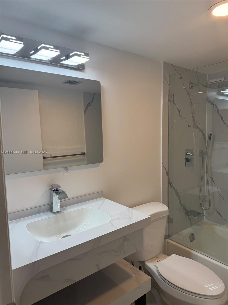 Bathroom featuring a vanity with stone-finish countertop and integrated sink, modern rectangular chrome faucet, wall-mounted mirror, tiered chrome light fixture, and a glass-enclosed shower with floor-to-ceiling veined stone-finish tile surround