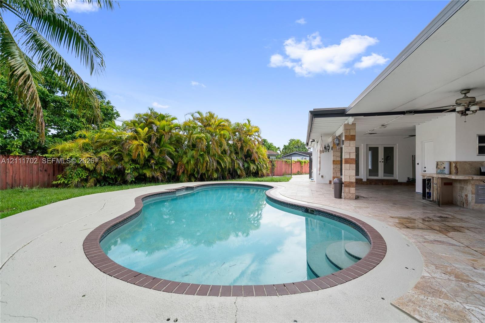 Free-form swimming pool featuring a brick-tiled coping and integrated stairs, expansive covered patio with tile flooring, built-in outdoor stone-finish kitchen, and privacy fencing