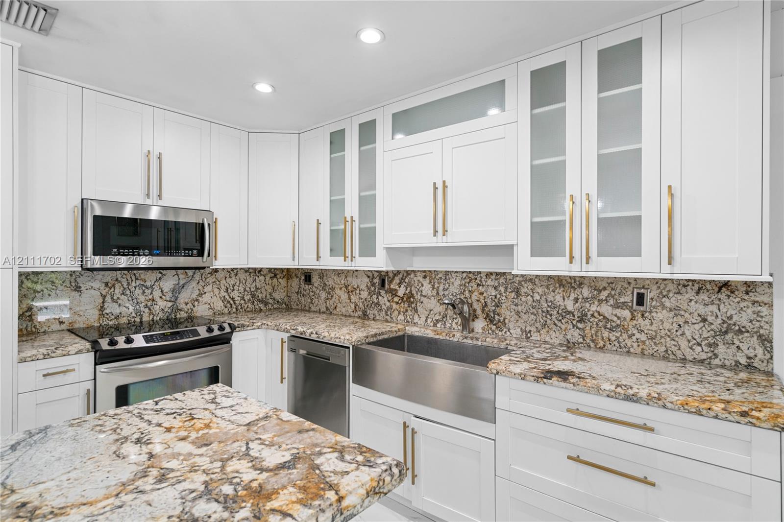 Kitchen featuring white shaker-style cabinetry with glass-panel uppers, stone countertops, stone backsplash, stainless steel farmhouse sink, and gold-tone hardware
