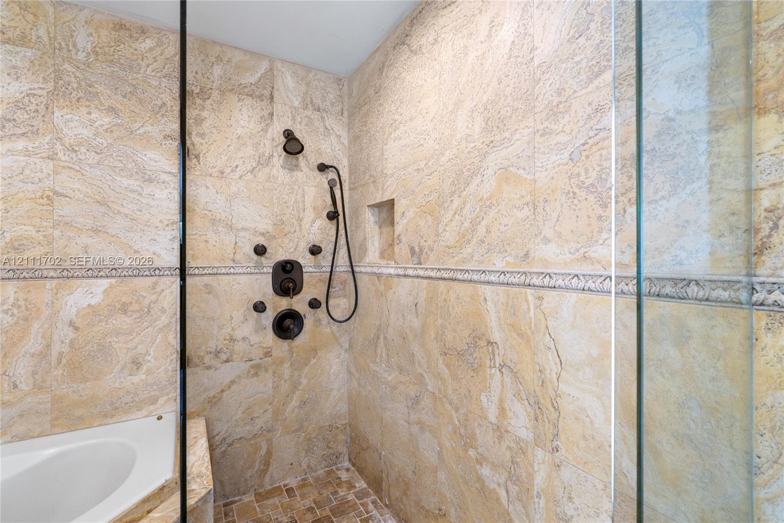 Walk-in shower featuring floor-to-ceiling stone tile, a mosaic tile inlay border, a multi-head shower system with a handheld sprayer, and an integrated wall niche