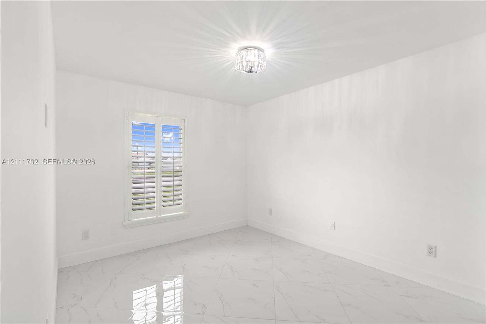 Polished stone-finish tile flooring, white walls, multi-pane window with white plantation shutters, and a crystal-accented flush-mount ceiling light