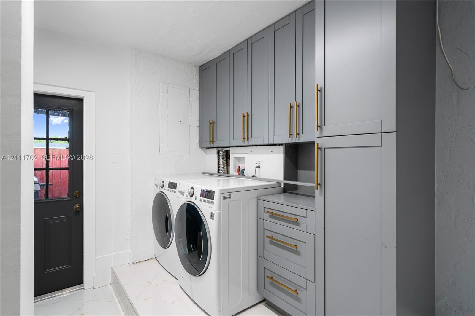 Laundry area featuring grey Shaker-style cabinetry, matte gold-finish hardware, and a dark-toned exterior door with glass pane detail