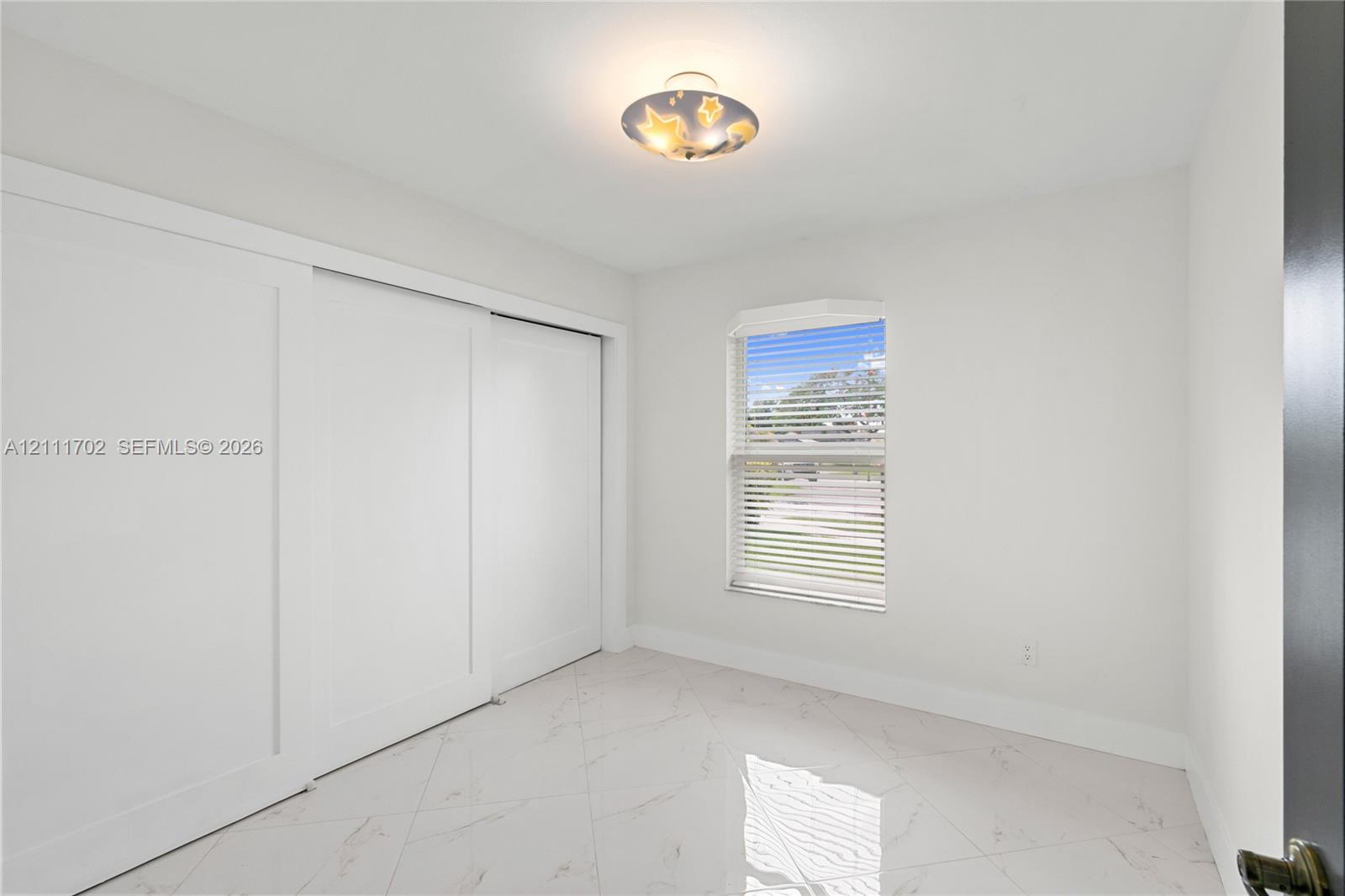 Room with polished tile flooring, sliding-door closet, single-pane window with blinds, and flush-mount ceiling light fixture