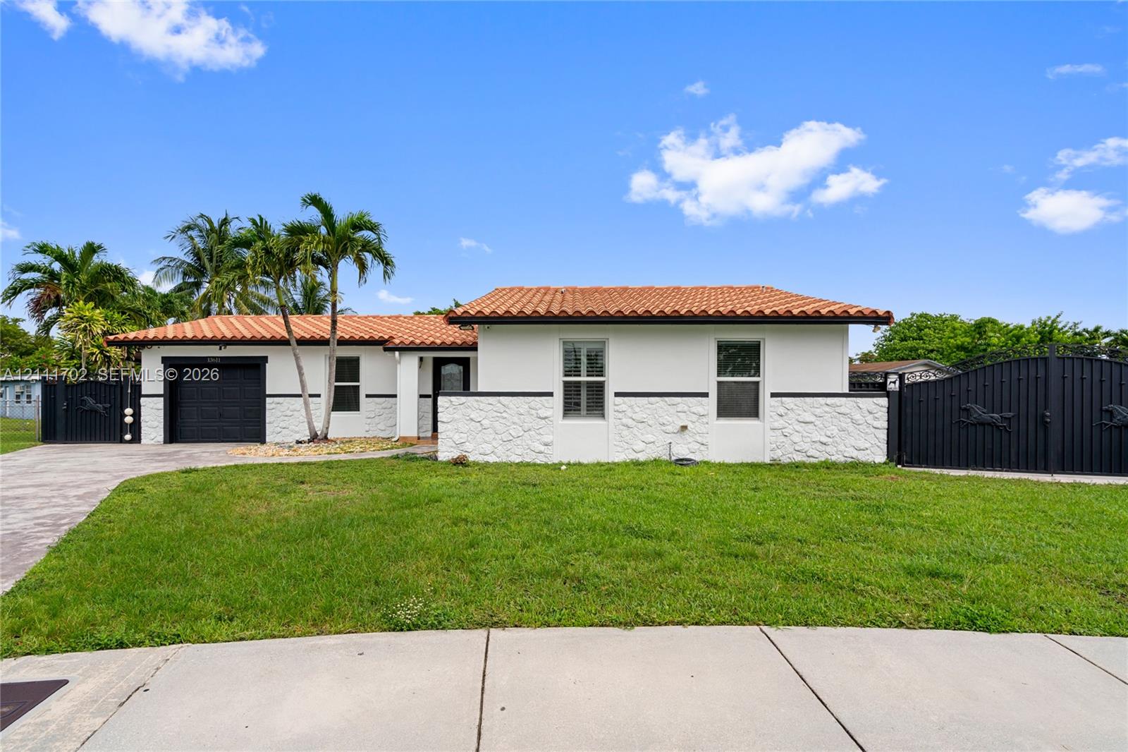 Single-story exterior featuring white stucco walls with stone veneer siding, terracotta tile roof, attached single-car garage, and custom black metal privacy gates