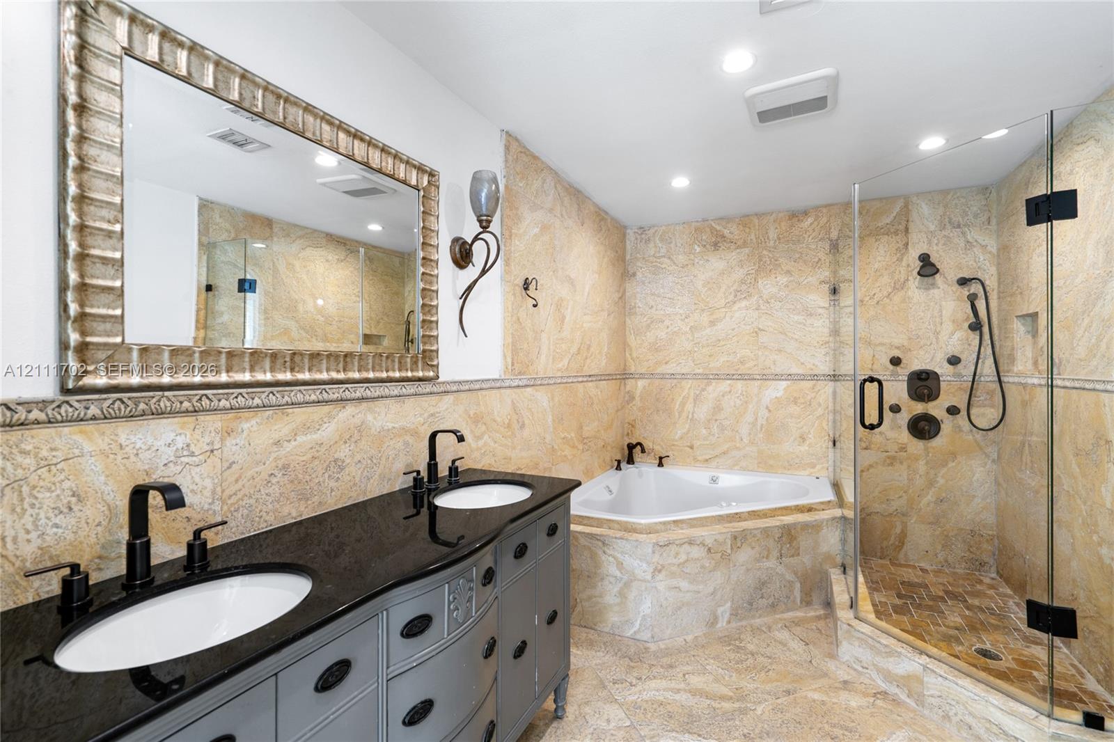 Dual-sink vanity with dark stone countertop, recessed lighting, corner jetted tub, and glass-enclosed walk-in shower featuring wall-mounted body sprayers and stone tile surround