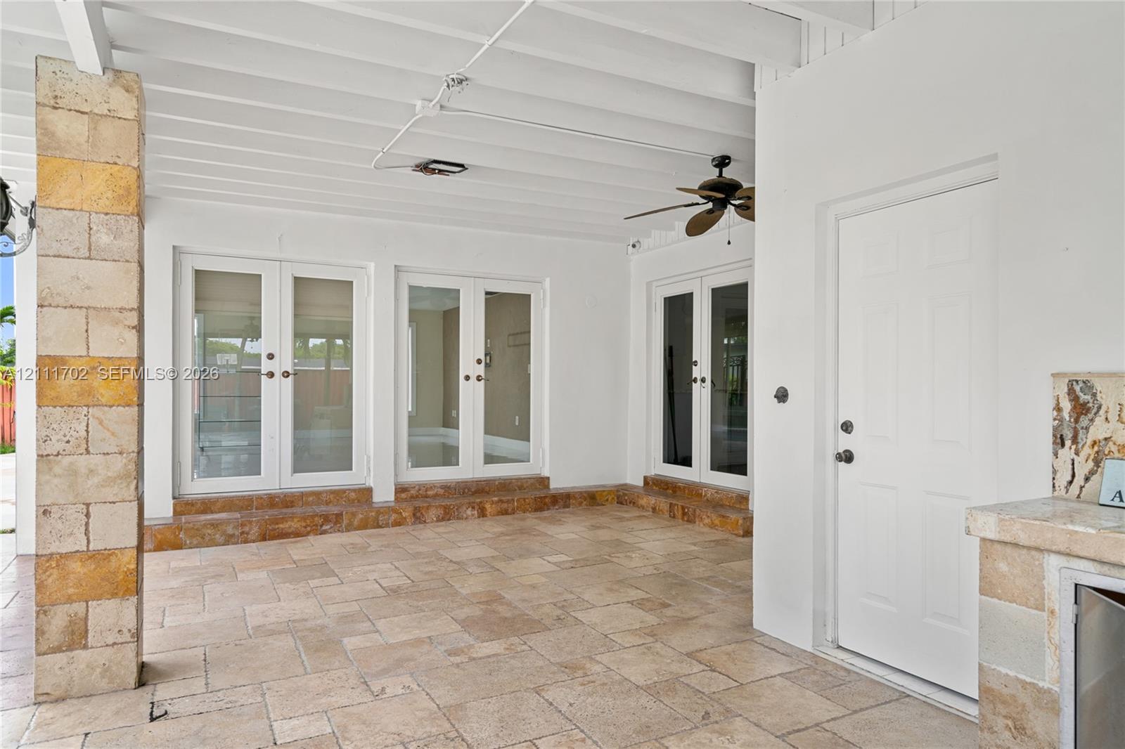 Covered patio featuring stone tile flooring, three sets of French doors, an outdoor kitchen structure with stone veneer, and a ceiling-mounted fan