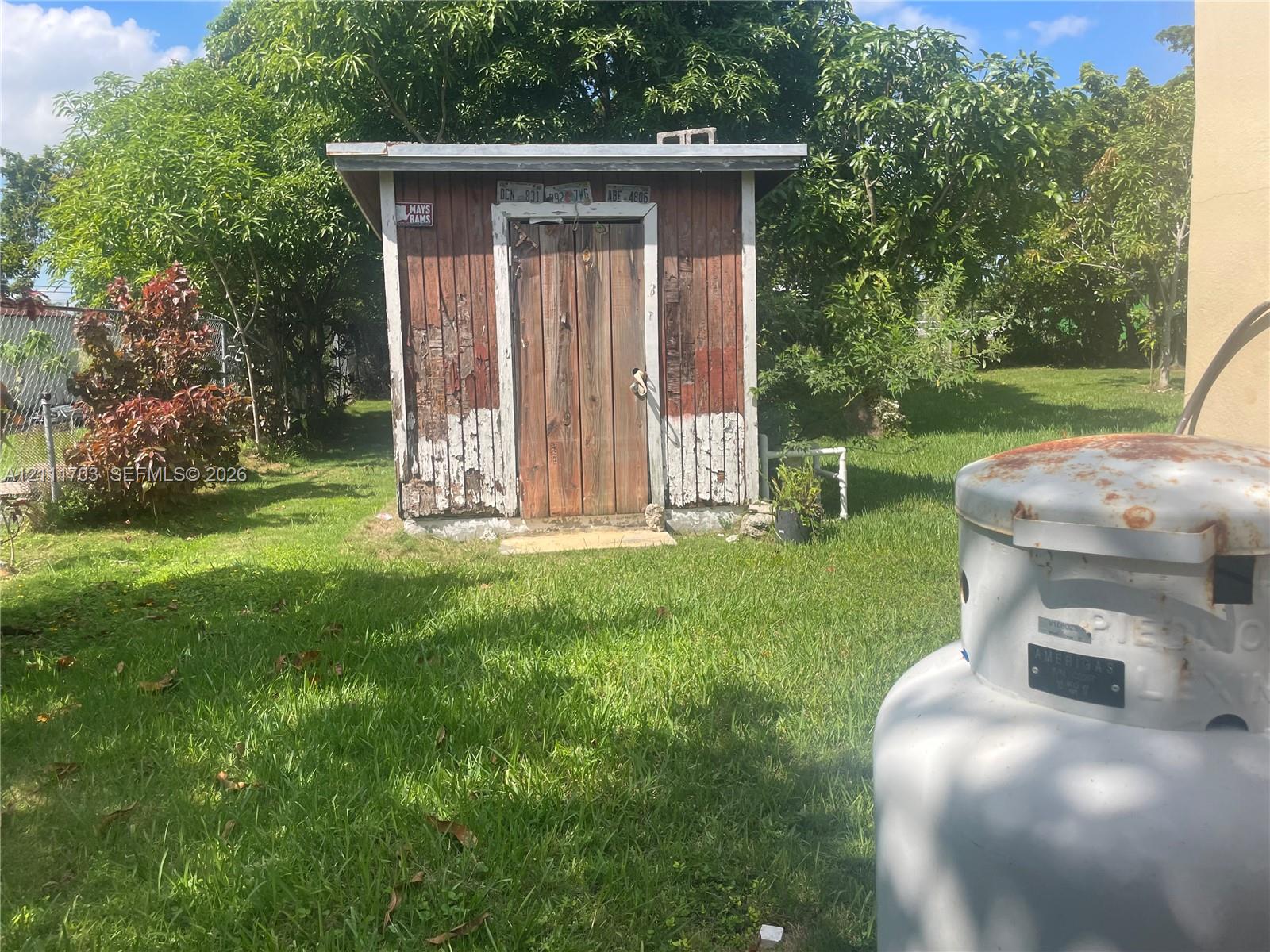 Outdoor storage shed with wood-paneled exterior and single door, situated on a grass-covered lot with mature trees