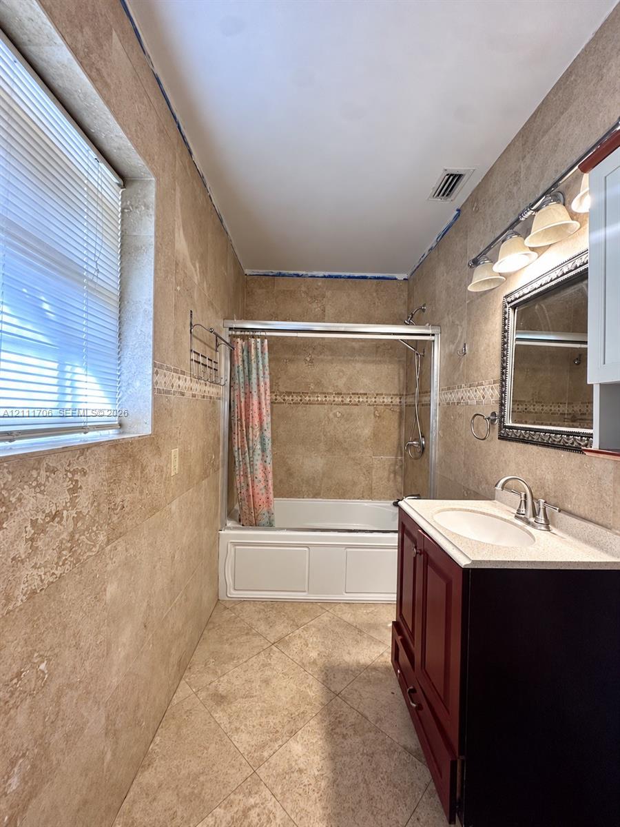 Full-height stone wall tiling with decorative inlay, walk-in bathtub with sliding glass doors, dark wood-finish vanity, light-toned stone countertop with an integrated sink, and wall-mounted vanity lighting