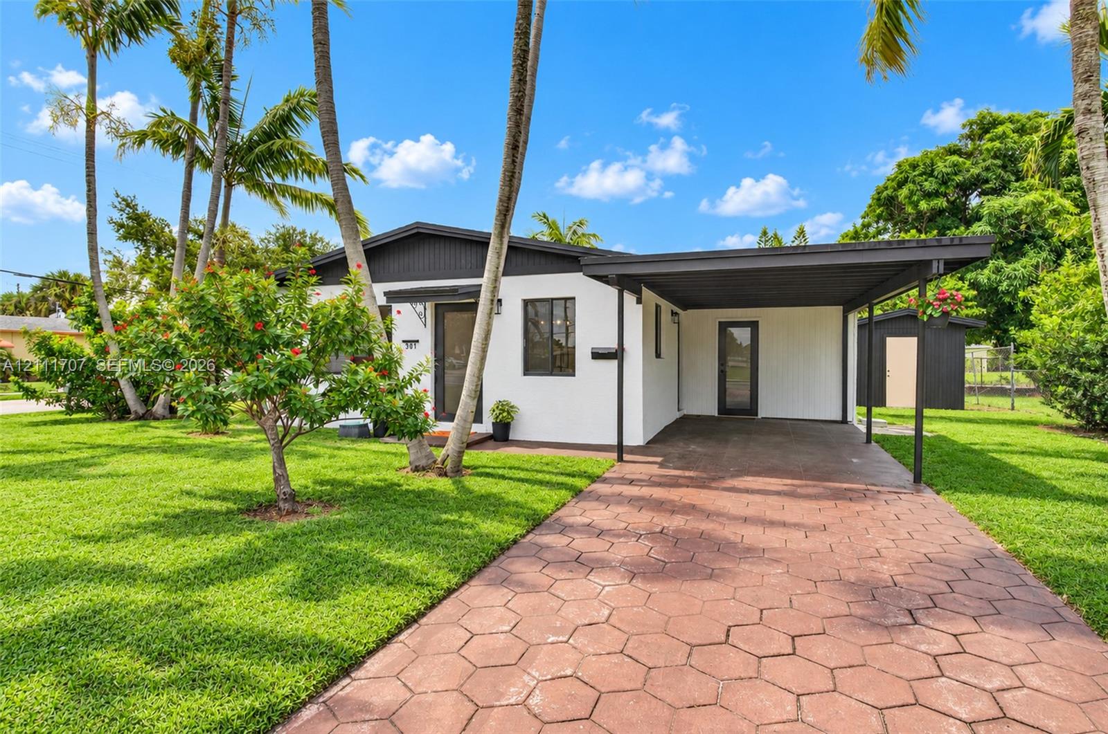 Single-story exterior featuring white stucco siding, a covered carport, hexagonal paver driveway, dark-toned trim, and established palm trees