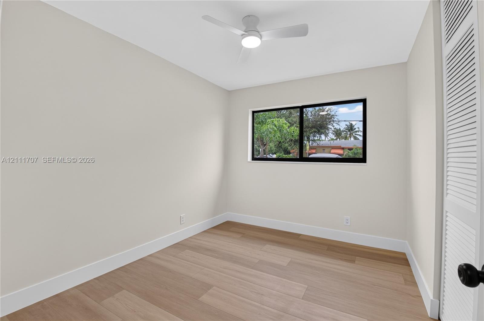 Neutral-toned interior featuring wood-finish flooring, a white ceiling fan with integrated lighting, a sliding window with a black frame, and a louvered bi-fold closet door