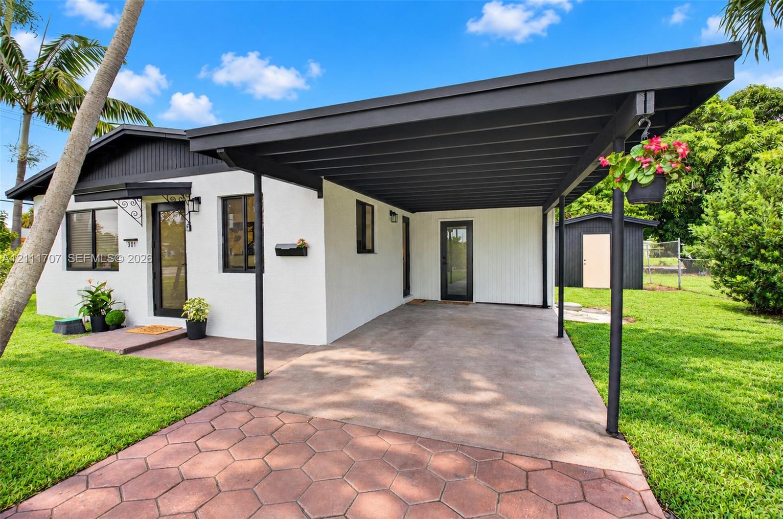 Exterior featuring a white stucco facade, covered carport with dark-toned trim, hexagonal paver driveway, and detached storage shed