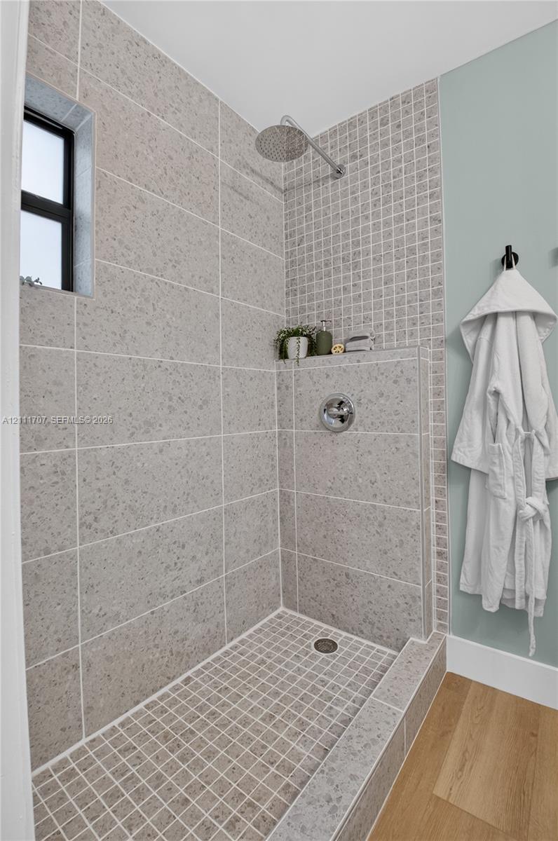 Walk-in shower featuring large-format neutral tile walls, a mosaic tile shower floor and accent wall, a built-in recessed shelf, a wall-mounted rainfall showerhead, and wood-finish flooring
