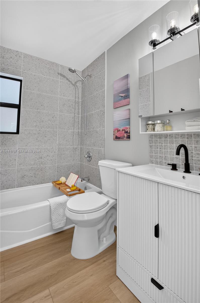 Bathroom featuring a ribbed-detail vanity with a matte black faucet, wood-finish flooring, a tub-shower combination with full-height stone-look tile surround, and a tri-light vanity fixture