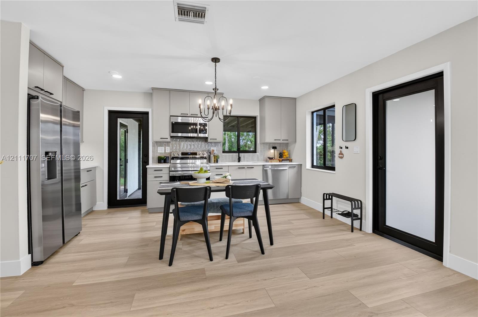 Kitchen featuring light-toned beige cabinetry, stainless steel appliances, wood-finish flooring, two exterior doors with black frames, and a multi-light chandelier