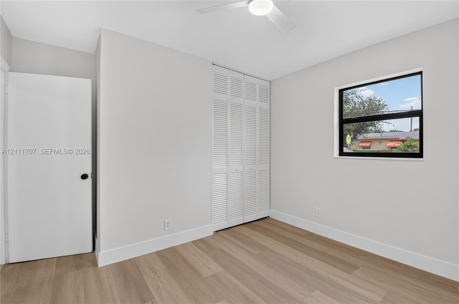 Room featuring wood-finish flooring, white louvered closet doors, a ceiling fan, and a window with a black frame