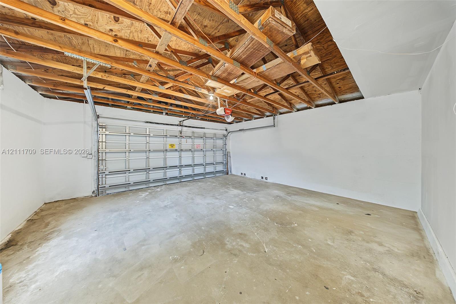 Garage interior featuring exposed rafter framing, white-painted walls, concrete flooring, and a sectional metal garage door