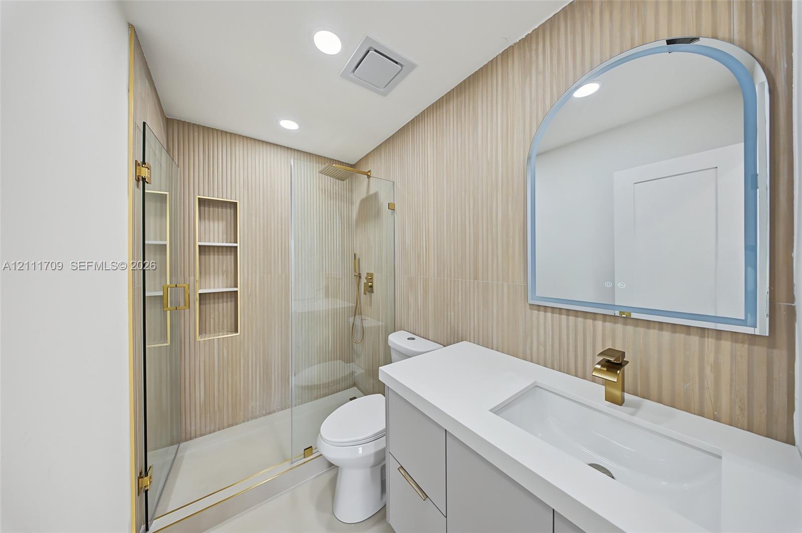 Bathroom featuring a frameless glass walk-in shower with integrated shelving, vertical-ribbed wall tile, a vanity with a white countertop and gold-finish faucet, and an arched wall-mounted mirror