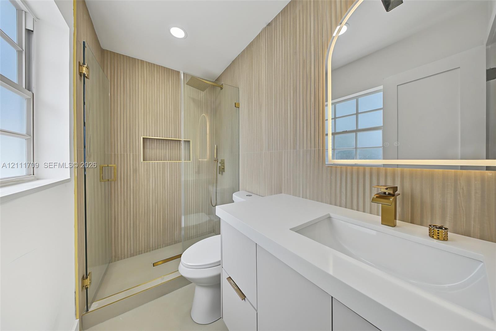 Bathroom featuring white vanity with integrated basin, gold-toned faucet, arched LED vanity mirror, vertical textured wall tile, and walk-in shower with glass enclosure