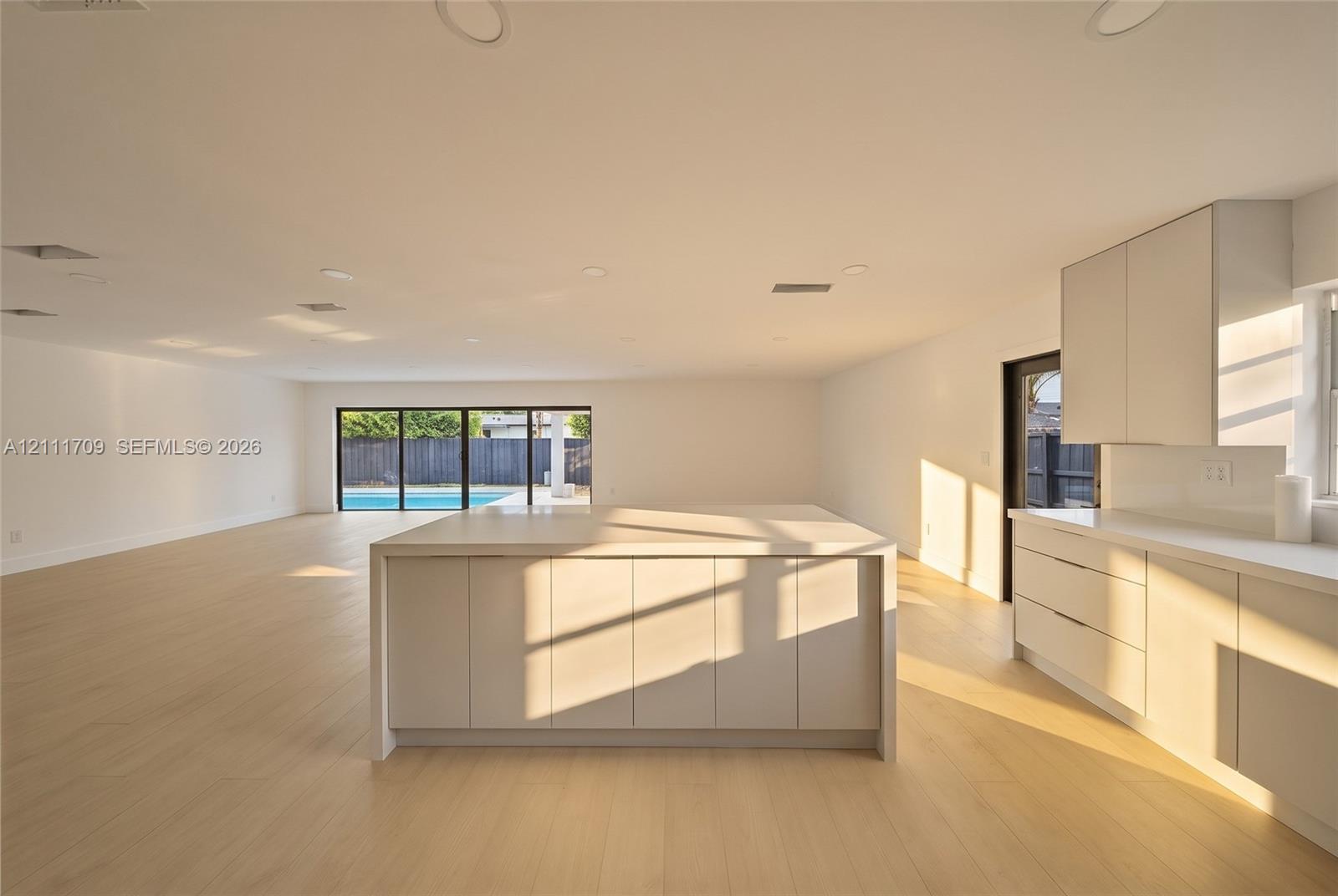 Open-concept interior featuring light-toned wood-finish flooring, a central kitchen island with integrated cabinetry, matte finish flat-panel cabinetry, and multi-panel sliding glass doors