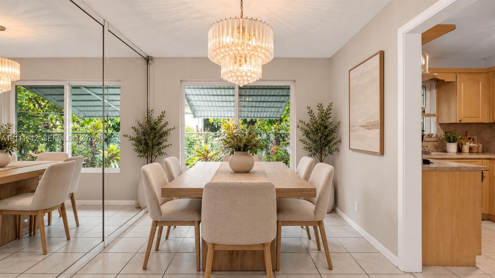 Virtually staged. Dining area featuring a tiered crystal chandelier, expansive wall-to-wall mirror paneling, neutral tile flooring, and large windows with outdoor awnings