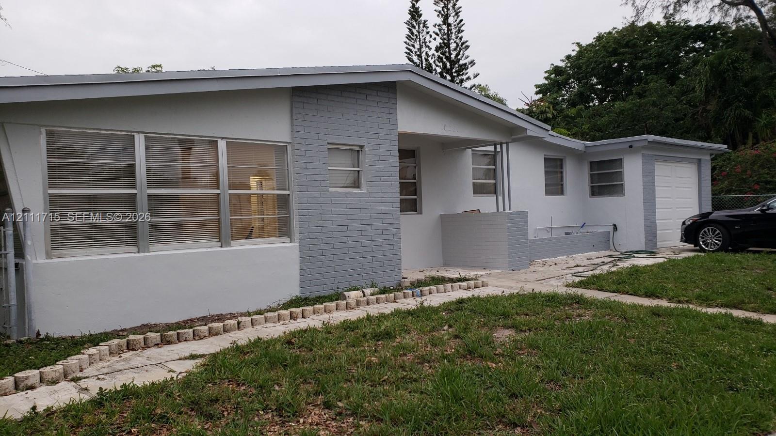Single-story exterior featuring painted brick accents, multi-pane horizontal windows, an attached garage, and a stone-bordered concrete walkway Single-story exterior featuring painted brick accents, multi-pane horizontal windows, an attached garage, and a stone-bordered concrete walkway