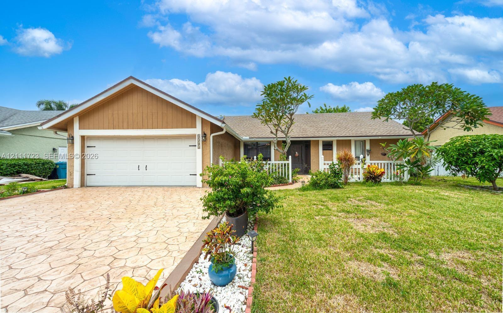 Single-story exterior featuring a stamped concrete driveway, two-car garage, white picket fencing, and mature landscaping